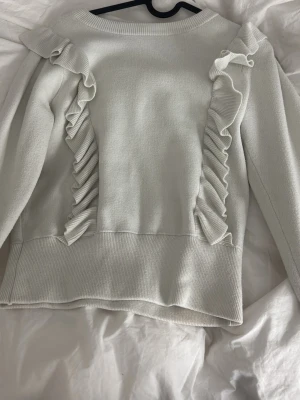 Vit stickad tröja med volangdetaljer - Snygg vit stickad tröja med långa ärmar och dekorativa volanger längs framsidan och axlarna. Tröjan har en rak passform och ribbstickade muddar vid nederkant och ärmslut. Perfekt för dig som gillar en söt och trendig look.