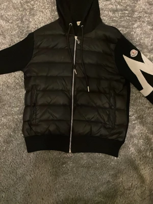 Svart pufferjacka med huva från Moncler - Snygg svart pufferjacka från Moncler med huva och dragkedja framtill. Jackan har vadderad kropp, stickade ärmar med Moncler-logga på ena ärmen och ribbade muddar. Perfekt för kalla dagar och riktigt trendig stil.