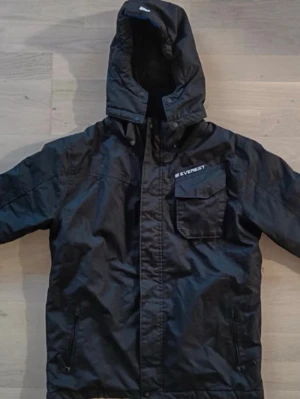 Svart parkasjacka från Everest - Svart parkasjacka från Everest med huva och dragkedja framtill. Jackan har en bröstficka med lock och två sidofickor med dragkedja. Materialet är slitstarkt och jackan har en klassisk, rak passform. Perfekt för kalla dagar.