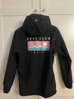Svart skid- och snowboardjacka från Dope Snow - Snygg och funktionell svart skid- och snowboardjacka från Dope Snow. Väldigt sparsamt använd och inga tecken på slitage!🤍                                          •Huva med dragsko.                               •Mediaficka                                                        •Tryck framtill + stort tryck på ryggen          •Mjukt och slitstarkt material                         •Resår vid ärmslut och nederkant            •Praktiska fickor med dragkedja + ficka till liftkort