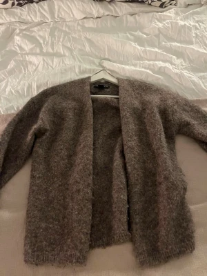 Brun fluffig kofta i mjukt material - Mysig brun kofta med öppen front och lång ärm. Koftan är fluffig och har en avslappnad passform, perfekt att slänga på sig när det är kyligt. Tillverkad i ett mjukt och värmande material som känns skönt mot huden.