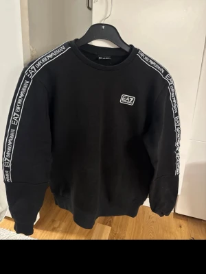 Svart EA7 Emporio Armani sweatshirt - Svart sweatshirt från EA7 Emporio Armani med logga på bröstet och tydliga logoband längs axlarna. Tröjan har rund halsringning och långa ärmar. Perfekt för dig som gillar stilrena och sportiga plagg med premiumkänsla.
