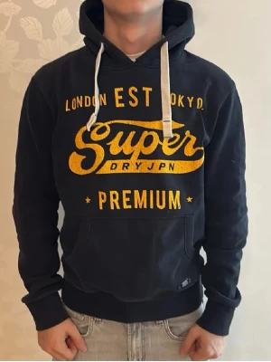 Svart Superdry hoodie med tryck - Snygg navy hoodie från Superdry med stort gult tryck på bröstet där det står 'Superdry Premium'. Hoodien har huva med vita snören och en klassisk känguruficka framtill. Perfekt för en avslappnad streetwear-look.