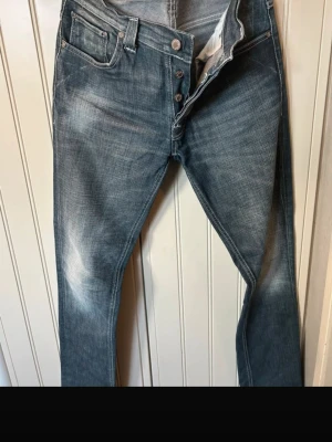 Blå jeans från Nudie Jeans - Säljer ett par klassiska blå jeans från Nudie Jeans med snyggt slitna detaljer och raka ben. Jeansen har knappgylf, fem fickor och kontrastsömmar på bakfickorna. Tillverkade i denim med en cool tvättad look.