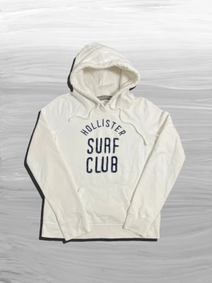 Hollister hoodie - Snygg vit hoodie från Hollister med broderad text 'Hollister Surf Club' i mörkblått på bröstet. Hoodien har huva med dragsko och en klassisk magficka. Tillverkad i mjukt bomullsmaterial, perfekt för chill dagar.