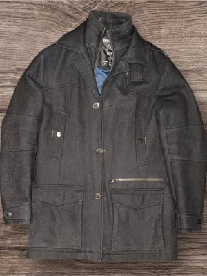 Svart kofta  - Stilren svart rock från spring Field med klassisk krage, flera fickor och knappstängning. Jackan har en blå insida och detaljer som dragkedja och knappar framtill. Perfekt för kyligare dagar och passar till många olika outfits.