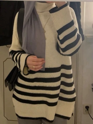 Randig stickad polotröja - Snygg stickad polotröja i vitt med svarta horisontella ränder. Tröjan har en lös passform, långa ärmar och hög krage. Perfekt för en trendig och avslappnad look.