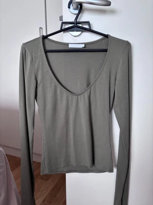 Olivgrön långärmad topp från A-DSGN - Snygg olivgrön långärmad topp från A-DSGN med djup v-ringning. Tunn och stretchig modell som sitter slimmat mot kroppen. Perfekt att styla med jeans eller kjol för en trendig look.Använd några gånger och sparsamt. Köpte den i 2025. 