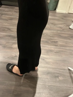 Svart ribbad långkjol - Snygg svart ribbad långkjol med slits bak. Kjolen har en tight passform och är perfekt för dig som gillar en stilren och enkel look. Materialet är stretchigt och följsamt, vilket gör den bekväm att bära hela dagen.