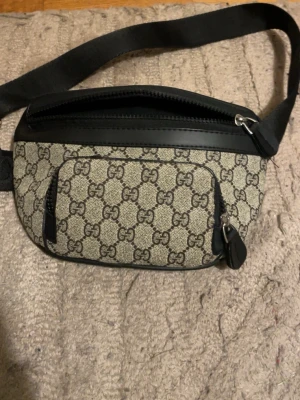 Gucci beige magväska med GG-mönster - Lådan får man ej med !!  Säljer en beige magväska från Gucci med klassiskt GG-mönster och svarta detaljer i läder. Väskan har justerbart band, två dragkedjefack och en stilren design som passar perfekt till streetwear eller casual outfits.