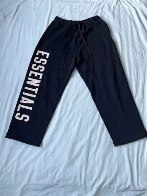 Svarta Essentials mjukisbyxor - Svarta mjukisbyxor från Essentials med stor vit logga längs ena benet. Byxorna har resår i midjan och dragsko för skön passform. Perfekta för en avslappnad och trendig stil. Materialet känns mjukt och bekvämt.