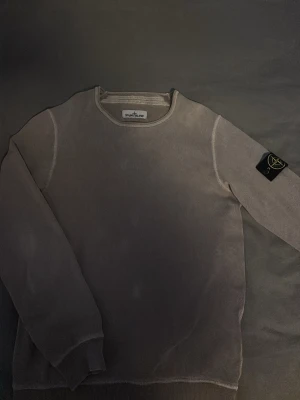 Grå Stone Island tröja - Säljer en stilren beige tröja från Stone Island med rund hals och den klassiska svarta logotypen med gult och grönt på vänster ärm. Tröjan har långa ärmarr. Perfekt för dig som gillar streetwear och enkel design. Små fläckar på tröjan men inga större problem. 