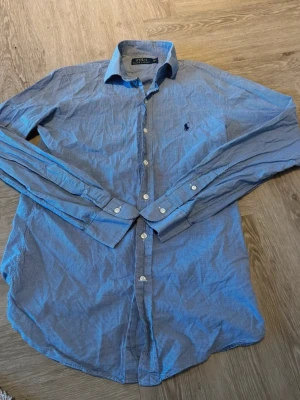 Blårutig skjorta från Polo Ralph Lauren - Klassisk blårutig skjorta från Polo Ralph Lauren med lång ärm och knappar framtill. Skjortan har en liten broderad logga på bröstet och är tillverkad i mjuk bomull. Perfekt för en stilren och avslappnad look.