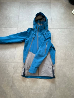 Oakley blå GORE-TEX vindjacka - Blå och grå vindjacka från Oakley i GORE-TEX Pro-material. Jackan har huva, vattentäta dragkedjor, flera fickor och justerbara ärmslut. Perfekt för dig som gillar outdoor och vill ha en sportig look. Oakley-logga på bröstet. Funkar kanon som en skid jacka med snölås