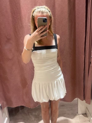 klänning från Gina ❤️ - Supersöt vit kort klänning med svarta axelband och fyrkantig urringning. Klänningen har en tight smockad överdel och en voluminös kjol med ballongformad nederdel