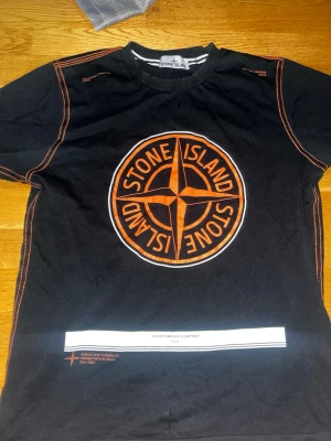 Svart Stone Island t-shirt med orange tryck - Snygg svart t-shirt från Stone Island med stort orange logotryck på bröstet och matchande orange sömmar längs axlar och sidor. Klassisk rund halsringning och korta ärmar. Perfekt för dig som vill sticka ut med streetwear-vibe.
