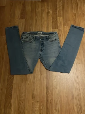 Ljusblå slim fit jeans från Jack N Jones - Säljer ett par ljusblå slim fit jeans från Jack N Jones med klassisk femficksdesign och dragkedjegylf. Jeansen har snygga slitningar och tvättade detaljer framtill och är tillverkade i stretchigt denimtyg för en bekväm passform.