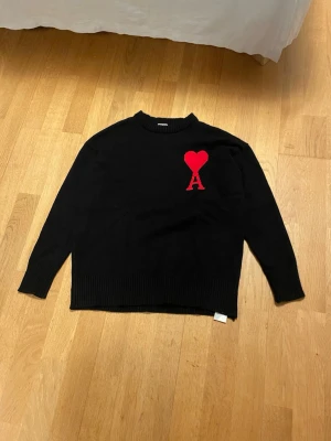 Svart stickad tröja från Ami Paris - Svart stickad tröja från Ami Paris med röd hjärtlogga och bokstaven A på bröstet. Tröjan har rund halsringning, långa ärmar och ribbstickade muddar. Perfekt för dig som gillar stilrena och trendiga plagg.