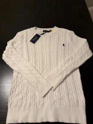 Vit kabelstickad tröja Polo Ralph Lauren - Kabelstickad vit tröja från Polo Ralph Lauren med rund halsringning och diskret broderad logga på bröstet. Långa ärmar och ribbade muddar ger en klassisk look. Perfekt för dig som gillar stilrena och tidlösa plagg.