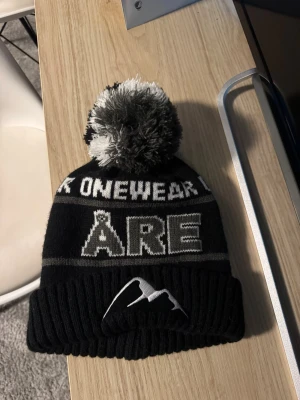 Svart Åre-mössa med tofs - Snygg svart mössa från OneWear med stor vit text 'ÅRE' framtill och en broderad bergslogga. Mössan har en svartvit tofs på toppen och ribbstickad kant. Perfekt för kalla dagar och vintersäsongen.