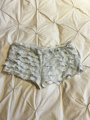 Grå spetsiga shorts med volanger - Supersöta grå shorts i spets med flera lager volanger och blommigt mönster. Shortsen har en mjuk resår i midjan och är tillverkade i ett stretchigt spetsmaterial som känns både bekvämt och snyggt.