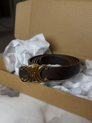 Brunt skärp från Celine med gulddetalj - Snyggt brunt skärp från Celine i äkta skinn med ikoniskt guldspänne i form av deras Triomphe-logo. Perfekt för att lyfta vilken outfit som helst med en exklusiv touch. Smal modell som passar både till jeans och klänning.