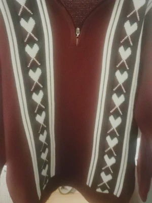 Vinröd stickad quarter zip med hjärtmönster - Vinröd stickad tröja med halv dragkedja och ståkrage. Tröjan har vita och bruna ränder samt hjärtmönster längs framsidan. Långa ärmar och ribbade muddar vid ärmslut och nederkant. Perfekt för kalla dagar och riktigt mysig stil.
