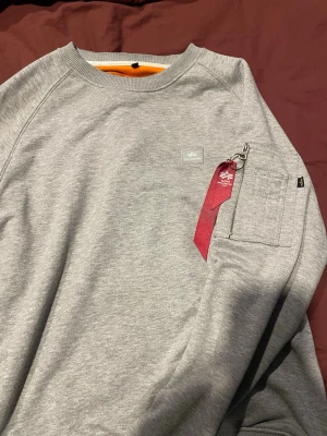 Grå sweatshirt Alpha Industries - Snygg grå sweatshirt från Alpha Industries med klassisk logga på bröstet och en unik ficka med dragkedja och röd tag på ärmen. Tröjan har rund halsringning och är tillverkad i mjuk bomullsmix för maximal komfort.