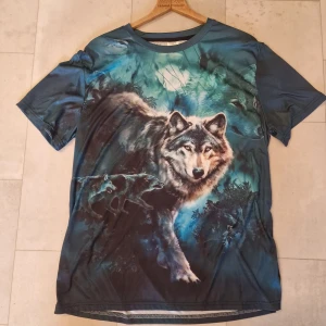 Blå t-shirt med vargtryck - Säljer en cool blå t-shirt med ett stort, detaljerat vargmotiv över hela framsidan och baksidan. T-shirten har korta ärmar och är gjord i ett mjukt, syntetiskt material som känns skönt mot huden. Perfekt för dig som gillar djur och unika prints.