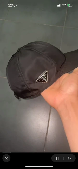 Svart nylonkeps från Prada - Snygg svart keps från Prada i glansigt nylonmaterial med klassisk böjd skärm och ikonisk metalltriangel med logga på sidan. Kepsen har paneldesign och justerbar rem baktill för perfekt passform. Perfekt accessoar för en trendig streetstyle.