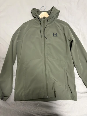 Olivgrön vindjacka från Under Armour - Snygg olivgrön vindjacka från Under Armour med huva och dragkedja framtill. Jackan har två stora fickor, meshfoder på insidan och justerbar dragsko vid huvan. Perfekt för blåsiga dagar och sportiga outfits.