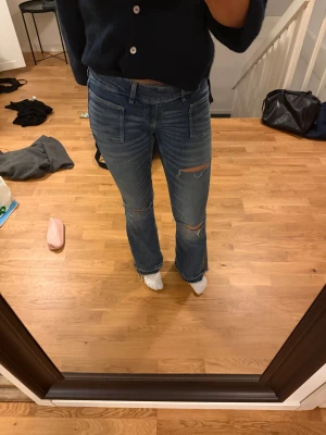 Blå bootcut jeans med slitningar - Säljer ett par blå bootcut jeans med låg midja och slitna detaljer vid knäna. Jeansen har fransad nederkant och stora fickor fram. Materialet är klassisk denim och passformen är loose med en snygg utsvängd siluett. Gina Young storlek m men skulle säga att det passar s 