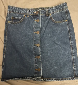 Mörkblå jeanskjol med knappar - Säljer en mörkblå jeanskjol med snygga kontrastsömmar och fem metallknappar framtill. Kjolen har klassiska fickor både fram och bak samt hällor för bälte. Modellen är rak och kort, perfekt till sneakers eller boots.