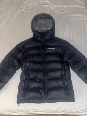 Svart Peak Performance jacka  - Säljer en svart dunjacka från Peak Performance med huva och dragkedja framtill. Jackan är vadderad i breda kanaler och har logga på bröstet. Perfekt för kalla dagar och har en clean, sportig look. Speciellt nu när det är kallt. 