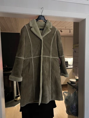 Mocka jacka från Saki, stl 46 - Säljer en lång mocka jacka från Saki med mjukt foder och markerade sömmar. Jackan har bred krage, uppvikta ärmslut och två fickor framtill. Perfekt för kalla dagar när du vill vara både varm och snygg.