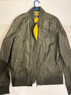 Olivgrön bomberjacka från J.Lindeberg - Snygg olivgrön bomberjacka från J.Lindeberg med gul insida. Jackan har ribbade muddar, två sidofickor med tryckknappar och en cool patch på bröstet. Dubbel dragkedja framtill och klassisk bomberkrage. Perfekt för dig som gillar stilren streetwear.