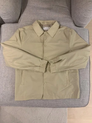 Beige coach-jacka från ASOS DESIGN - Snygg beige coach-acka från ASOS DESIGN i storlek S. Jackan har klassisk krage, raka ärmar och knäppning med tryckknappar framtill. Perfekt för dig som gillar stilrena och minimalistiska plagg. Materialet är lätt och känns skönt mot kroppen.