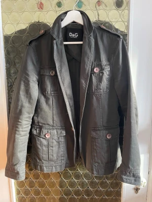 Mörkgrön kavajjacka från Dolce & Gabbana - Snygg grå kavajjacka från D&G med klassisk krage, axelklaffar och fyra framfickor med knappar. Jackan är tillverkad i bomull och har en stilren, rak passform som ger en cool och avslappnad vibe. Perfekt för dig som gillar italiensk design och detaljer.