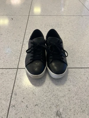 Svarta sneakers med vit sula - Snygga svarta sneakers med klassisk rund tå och vit platt sula. Skorna har snörning och är tillverkade i skinn för en stilren look. Perfekta för dig som gillar enkel och clean stil.