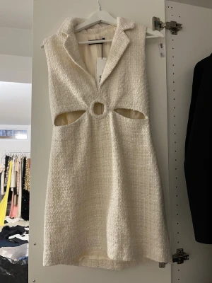 Vit kortklänning med cut-outs - Säljer en vit kortklänning från Zara med grov textur och snygga cut-outs vid midjan. Klänningen är ärmlös, har krage och ett cirkelformat hål framtill. Materialet känns som en blandning av bomull och syntet, perfekt för dig som gillar trendiga detaljer.