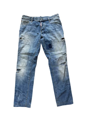 Dsquared2 blå slitna jeans - Säljer ett par blå jeans från Dsquared2 med coola slitningar, patchade detaljer och raka ben. Klassisk femficksmodell med Dsquared2-logga på röd etikett. Jeansen har en avslappnad vibe och är tillverkade i bomull med snygg tvättad look.