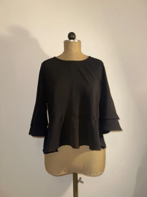 Svart blus med volangärmar - Säljer en svart blus med rund halsringning och volangdetaljer på de trekvarts långa ärmarna. Blusen har en lös passform och ett lätt rynkat parti nedtill som ger en snygg siluett. Perfekt för dig som gillar stilrena och trendiga plagg. ❣️