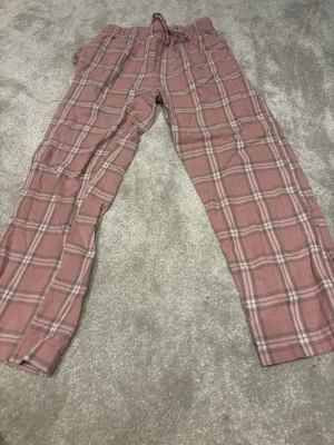 Rosa rutiga mjukisbyxor med snörning - Säljer ett par chill rosa mjukisbyxor från 157 med rutigt mönster i grått och vitt. Har en liten vit prick på högra låret men inget som syns. Byxorna har elastisk midja med snörning och är gjorda i mjukt bomullsmaterial. Perfekta för att hänga hemma eller mysa i soffan.