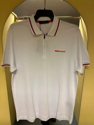 Vit polotopp från Prada med röda detaljer - Snygg vit polotopp från Prada med röda ränder på krage och ärmslut. Toppens klassiska design har korta ärmar och en diskret dragkedja framtill. Materialet är mjuk bomull och loggan syns på bröstet i rött. Perfekt för dig som gillar stilrena märkesplagg.