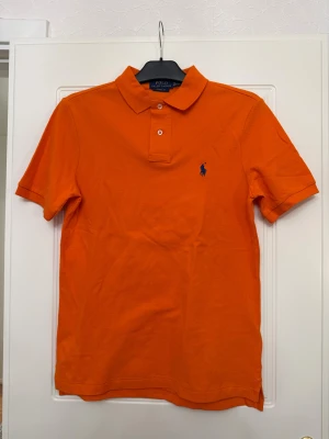 Orange piké från Polo Ralph Lauren - Snygg orange pikétröja från Polo Ralph Lauren med klassisk krage, två knappar och korta ärmar. Tröjan har det ikoniska blå broderade Polo-logot på bröstet och är gjord i mjuk bomull som känns skön mot huden. Perfekt för dig som gillar färg och stil.