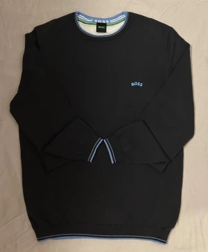 Mörkblå  tröja från BOSS med blå detaljer - Mörkblå  långärmad tröja från BOSS med rund halsringning och blå broderad BOSS-logga på bröstet. Tröjan har blå och vita ränder på muddar och krage för en clean look. Perfekt för dig som gillar stilrena och klassiska plagg.