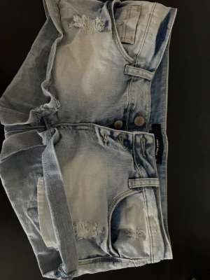 Ljusblå denimshorts med slitningar - Säljer ett par ljusblå denimshorts från H&M med snygga slitningar och upprullade benslut. Shortsen har klassisk femficksdesign, bälteshällor och dubbla knappar framtill. Perfekta för en chill sommarlook.