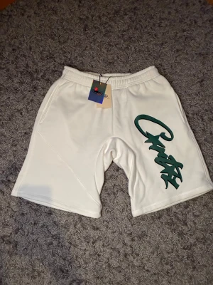 Vita shorts från Corteiz med tryck - Snygga vita shorts från Corteiz i storlek S med grönt graffiti-inspirerat tryck på ena benet. Modellen har elastisk midja och är tillverkad i mjukt syntetmaterial, perfekt för chill dagar. Klassisk loose fit och stilren design med fickor på sidorna.