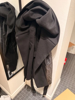 Svart kappa med stor huva och bälte - Säljer en svart kappa med stor, mysig huva och knytbälte i midjan. Jackan har lång ärm och stilren design, Passformen är normal och längden går till höften. Vid snabb affär kan jag gå ner i pris,