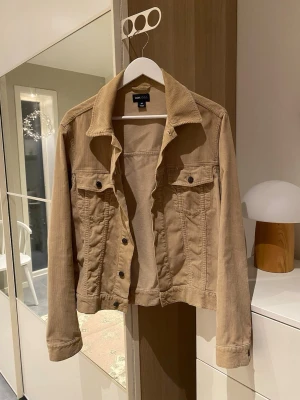 Beige manchester jeansjacka - Snygg beige jeansjacka från H&M i mjuk manchester. Klassisk modell med krage, knappar framtill och två bröstfickor med lock. Jackan har långa ärmar och en rak passform som funkar till många olika stilar. Storlek M
